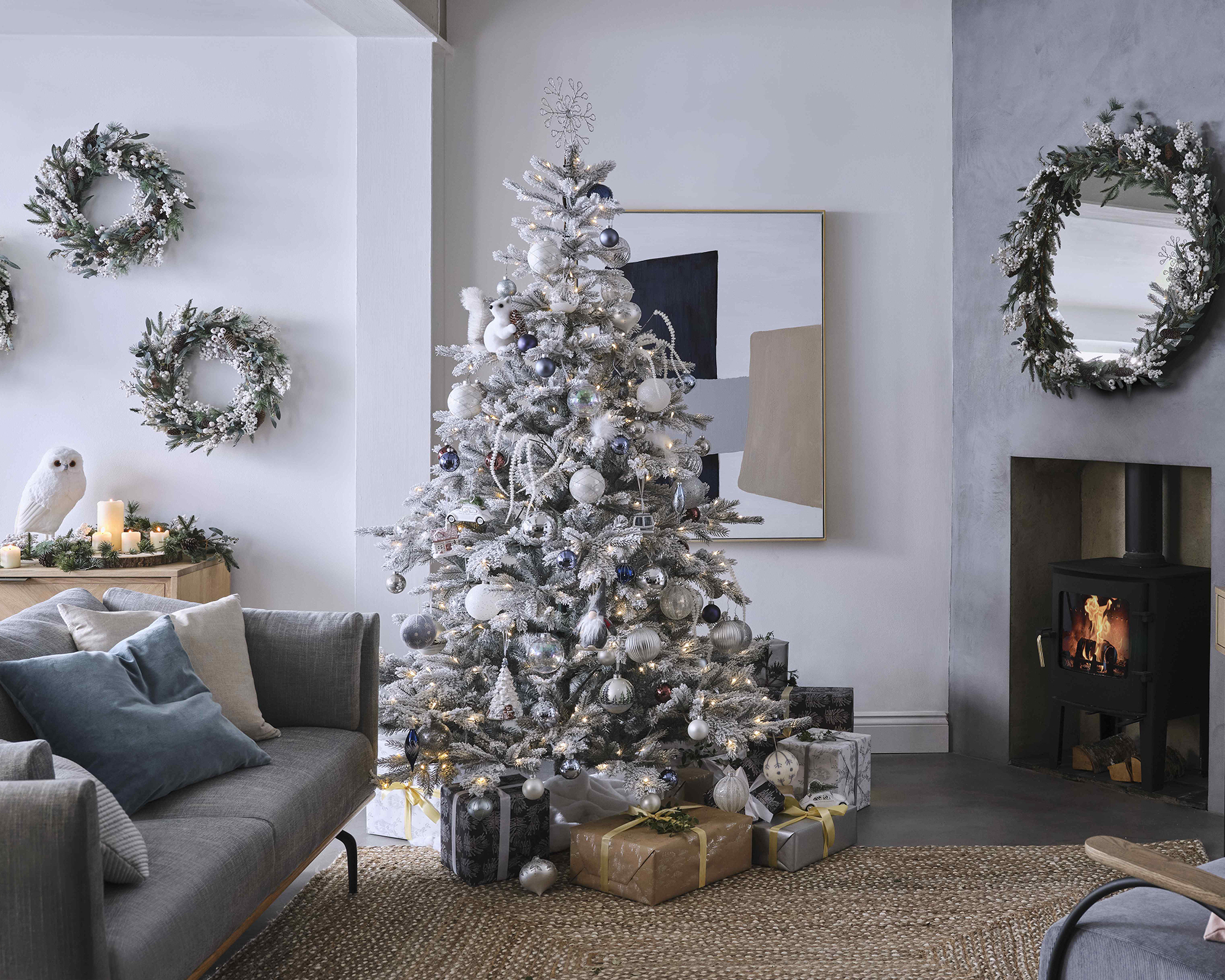 A Scandinavian-style grey and white living room with snowy faux Christmas tree, trio of wreaths on wall and traditional fireplace with flume