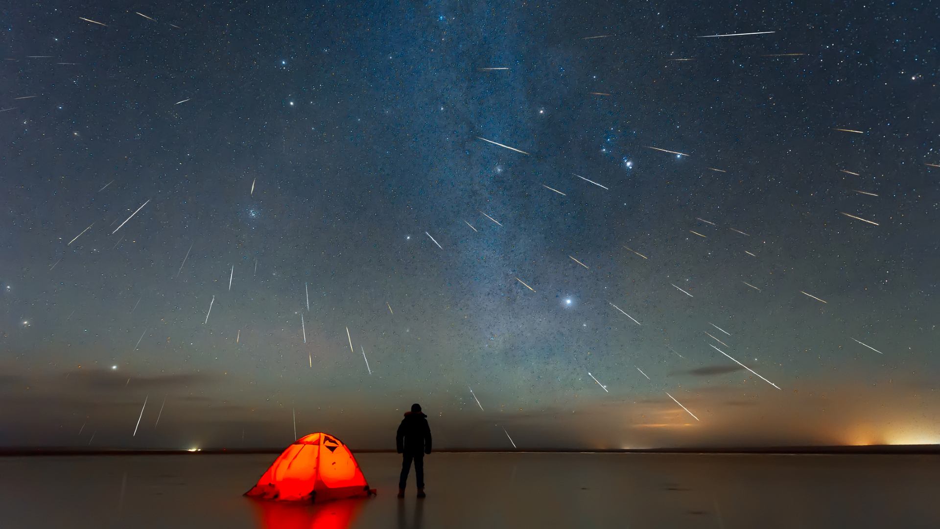A purple night sky filled with white streaks from Geminid meteors with a bright red tent and a person standing in the foreground