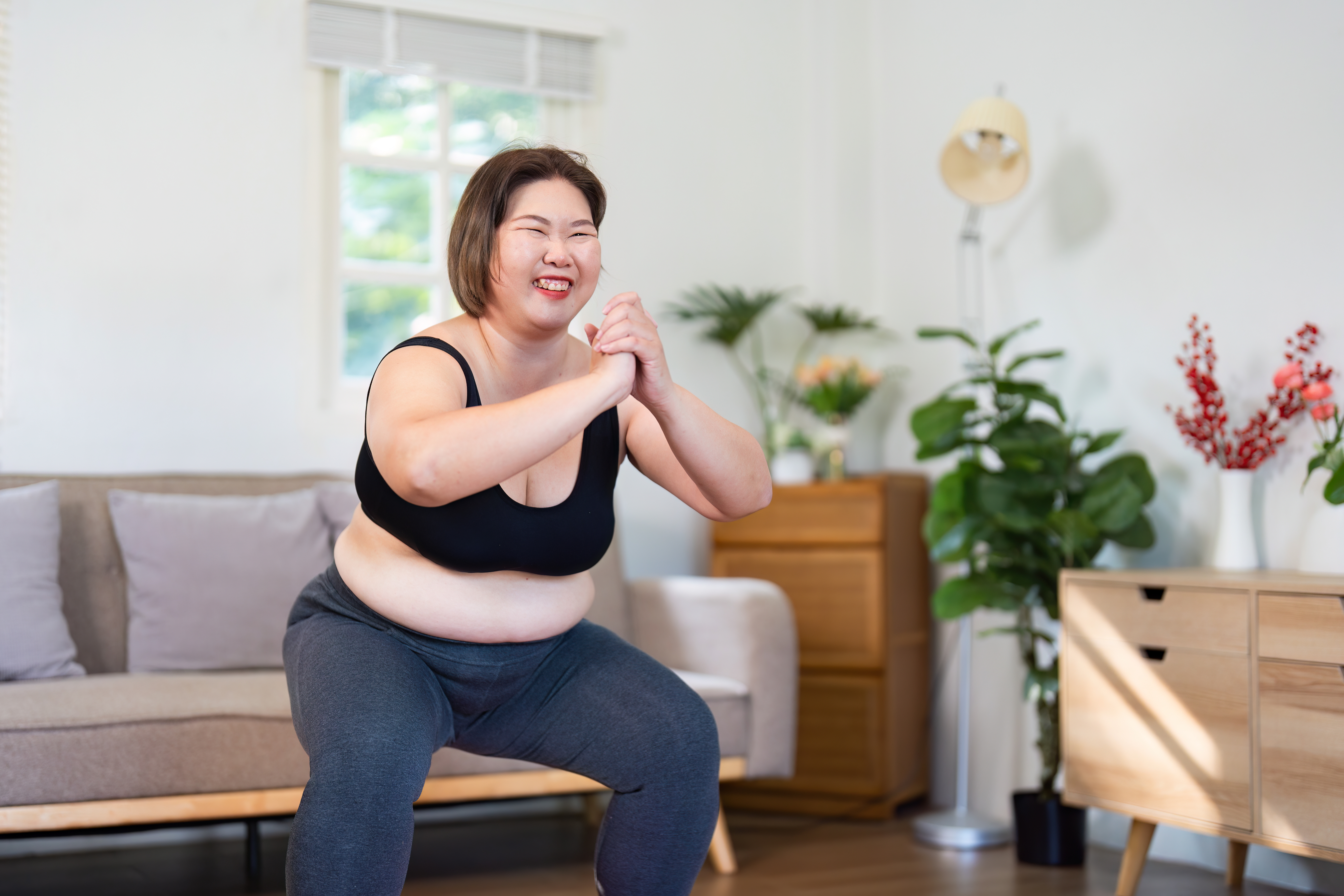 A woman smiles while working out at home in a sports bra and leggings. She is squatting, with her knees out wide and her hands clasped together. Behind her we see a couch and drawers.