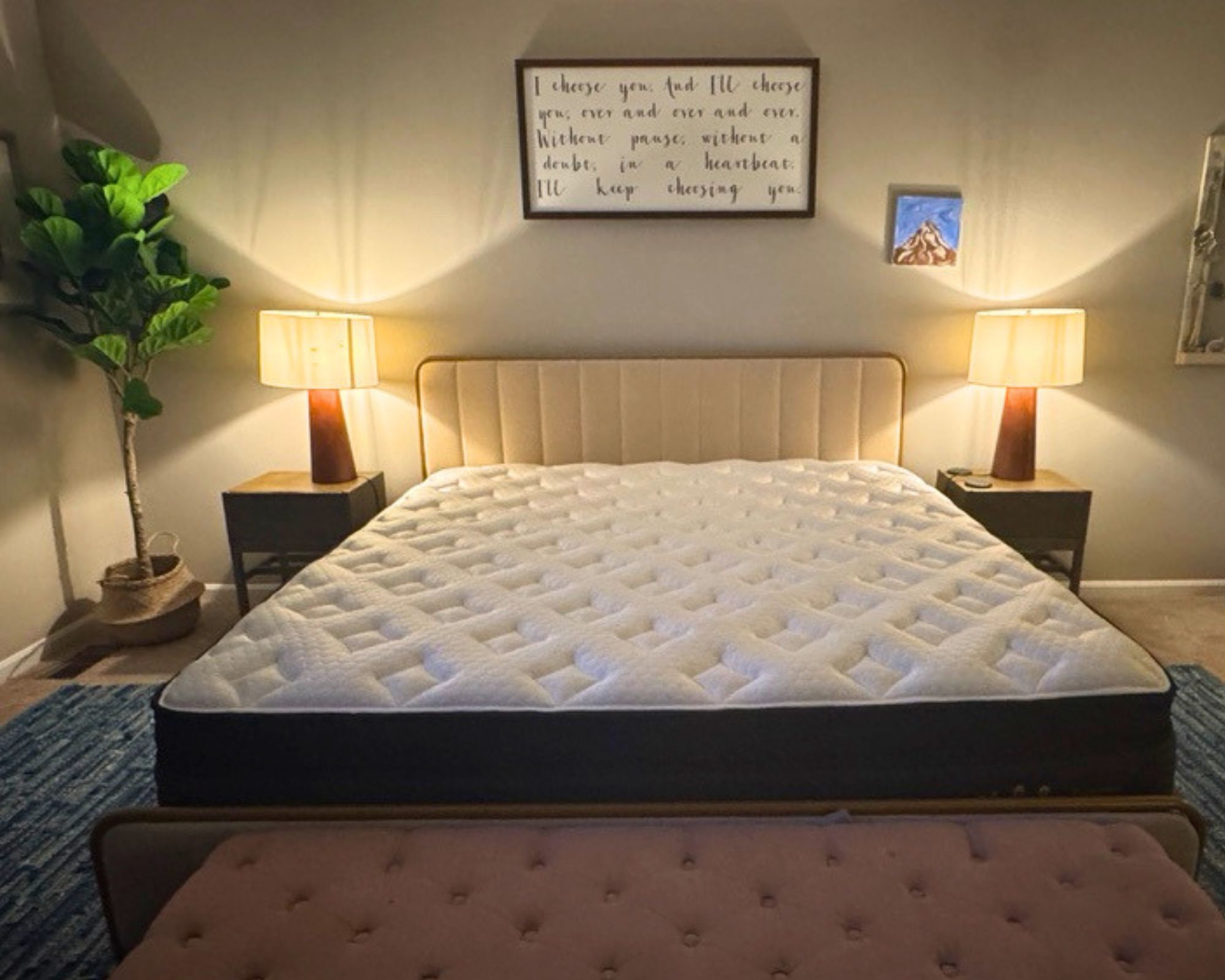 Nolah Evolution Mattress pictured in a white bedroom with a fluted headboard, two matching nightstands with red and white lamps, a large ottoman, houseplant, blue rug, and artworks hung on the wall.