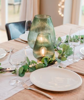 A pink dining table with a white runner, clear wine glasses, white circular plates, silver cutlery, and two green ridged vases in the middle and a eucalyptus garland