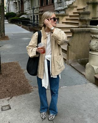 British fashion influencer Lucy Williams poses on a NYC sidewalk wearing black sunglasses, a barn coat, a black tote bag, a sweater tied around the waist, relaxed jeans, and snakeskin flats.