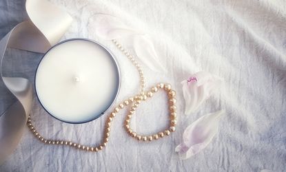 View Of Scented Candle And Pearl Jewelry With Ribbon On White Bed