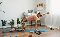 Woman on her hands and knees on the floor lifts a light yellow dumbbell in front of her. She is in a home scene, with plants and a desk in the background. 