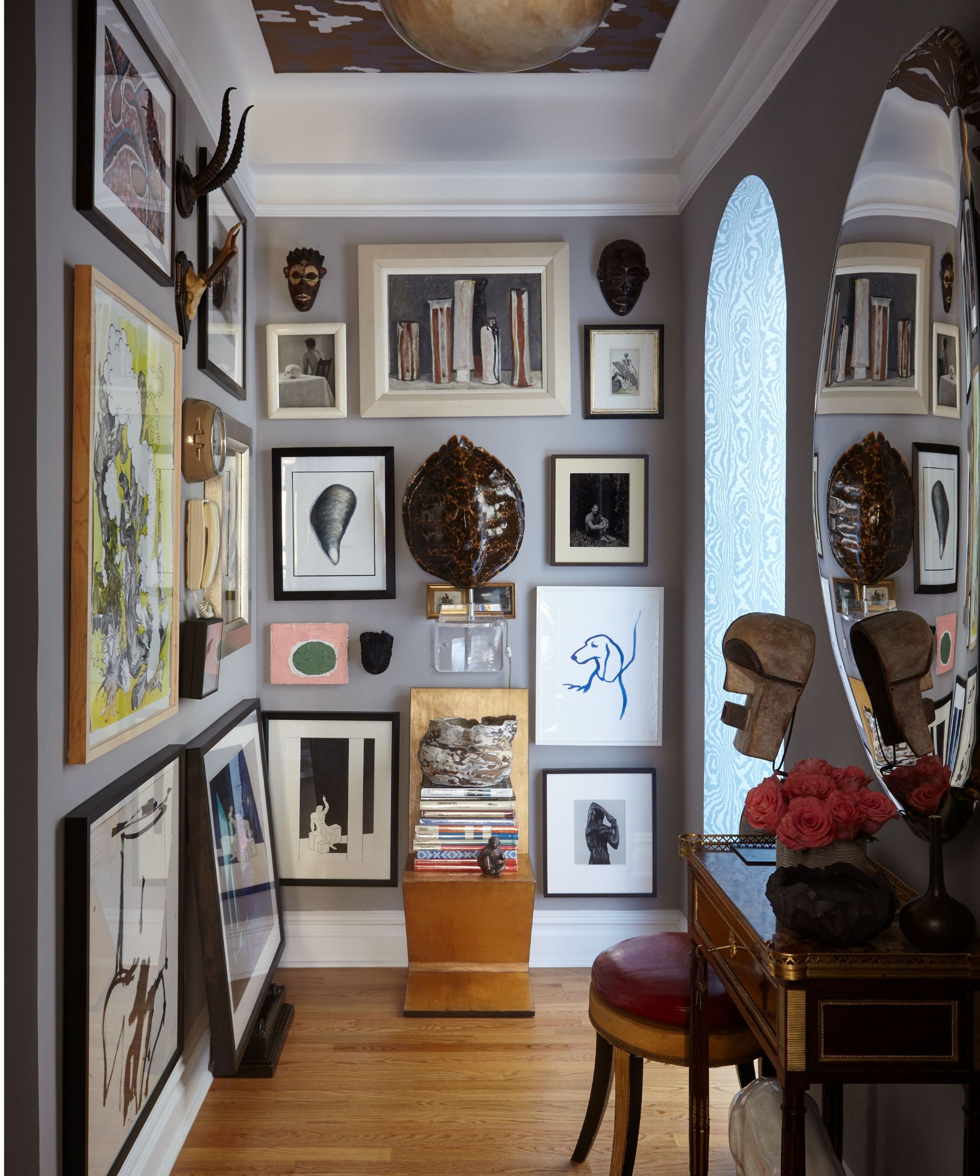 Entryway with pale blue walls, art on walls, arched doorway, wooden floor, antique console table and a leather red stool underneath