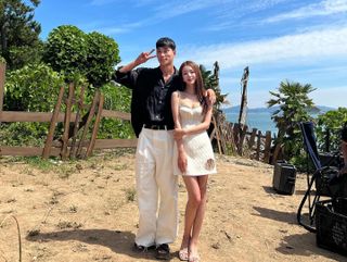 a man makes a v pose next to a woman holding her arm on a sandy tree-lined beach