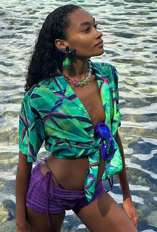 The shell jewelry trend shown on a woman wearing oversize starfish and shell earrings with a green tropical-print button-down shirt layered over a purple bikini and purple crochet shorts.