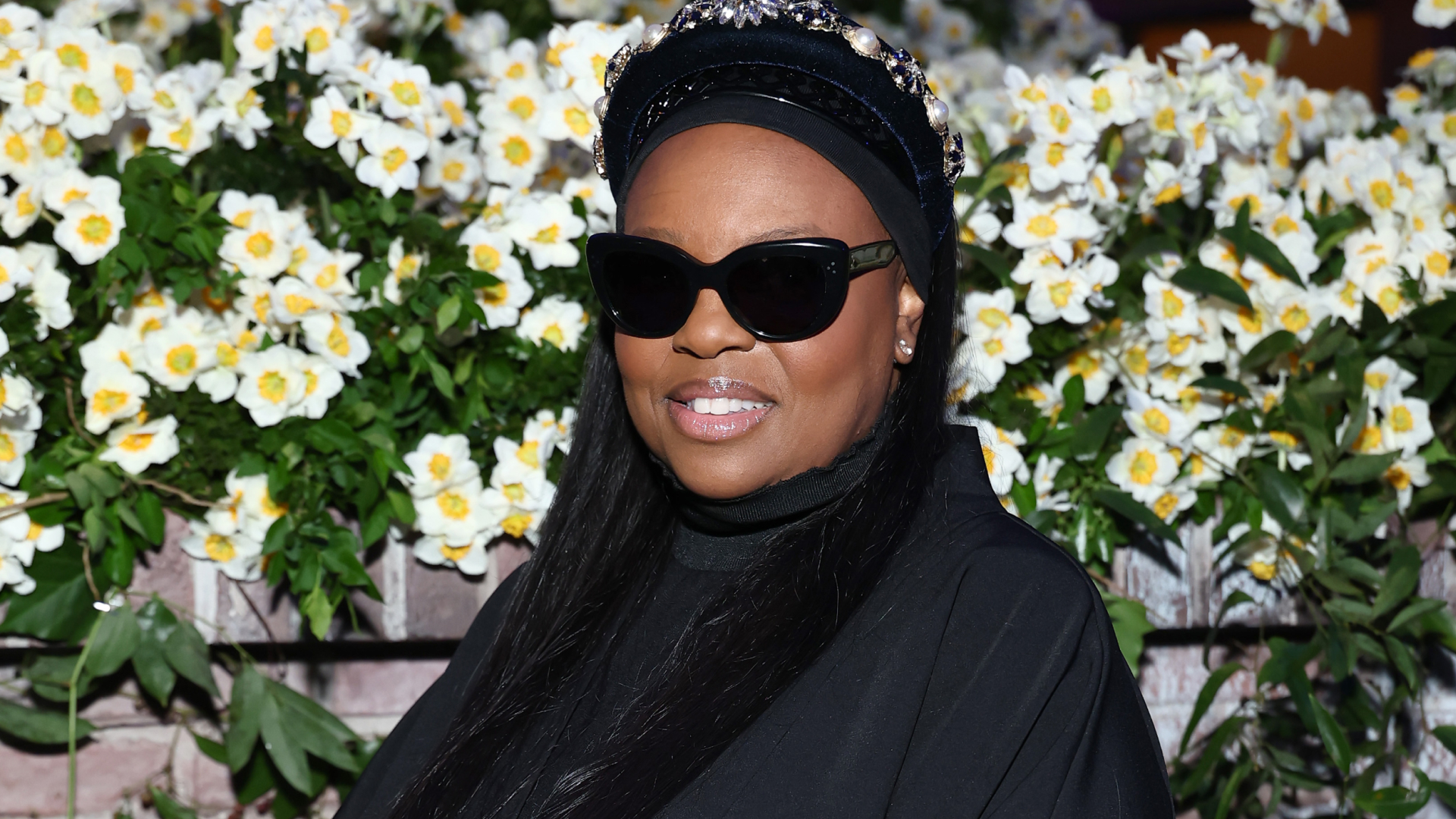 Dame Pat McGrath wearing sunglasses and a black turtleneck sweater. She is standing in front of a bouquet of flowers.