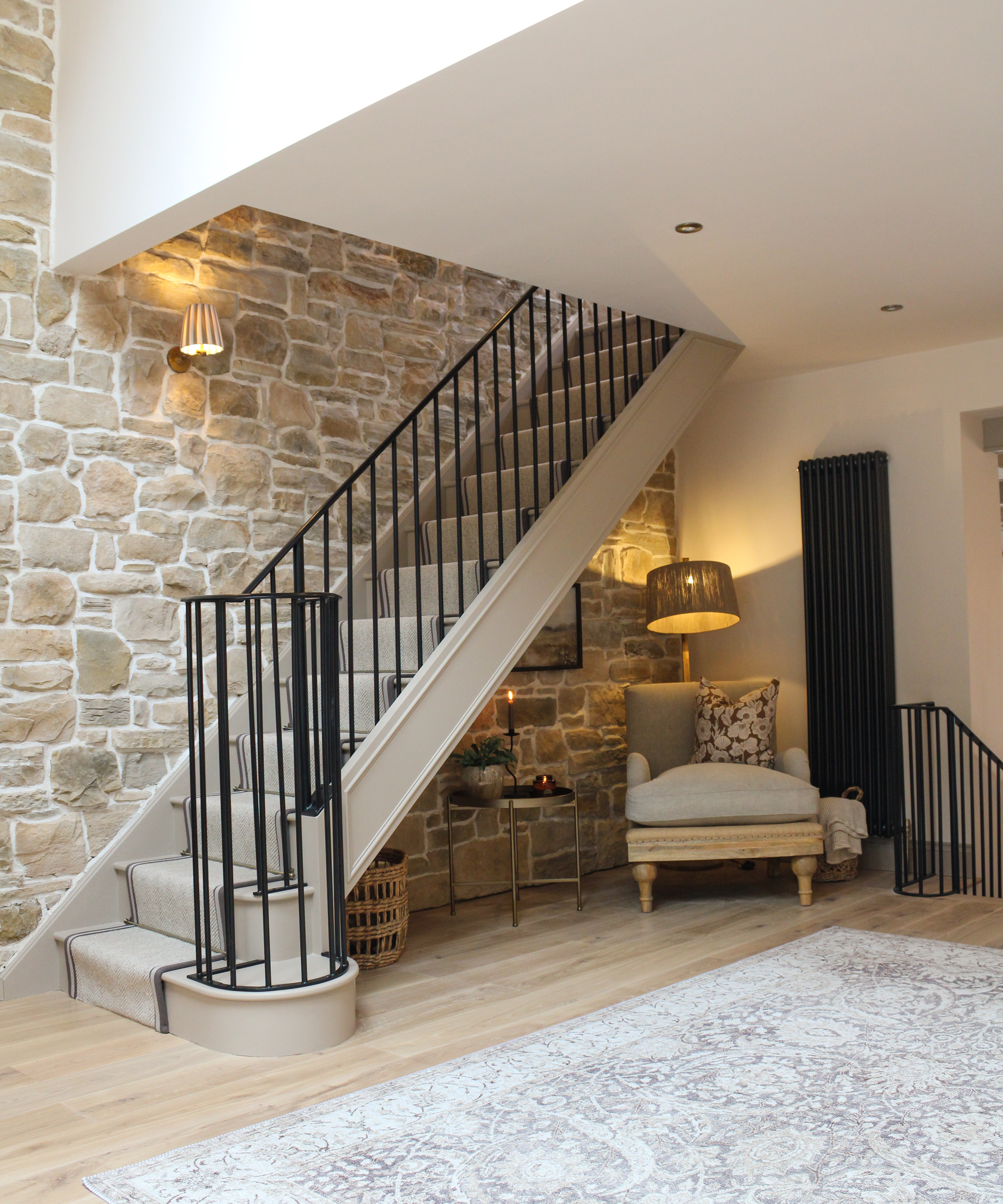 hallway with stone wall, wooden flooring, staircase with metal spindles, small wall light and floor light illuminating space below stairs