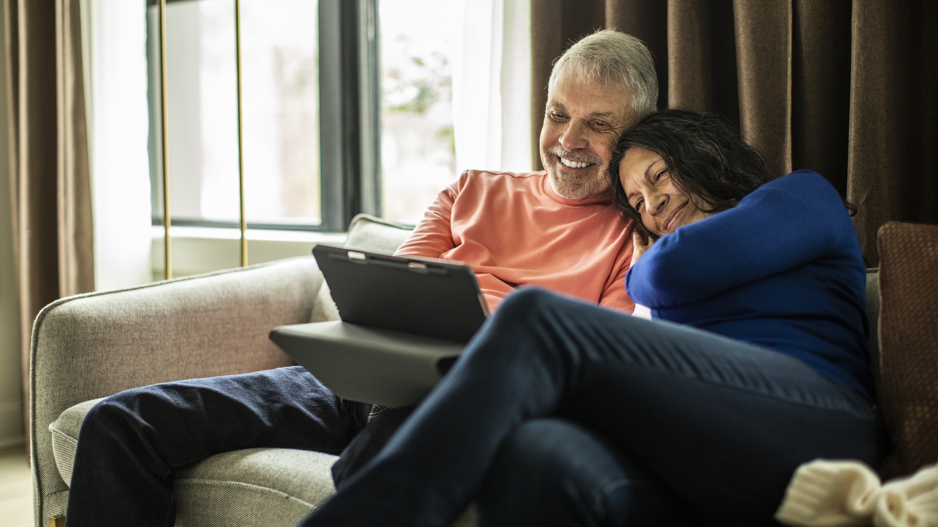 An older couple smile as they cuddle on their sofa and look at a tablet together.
