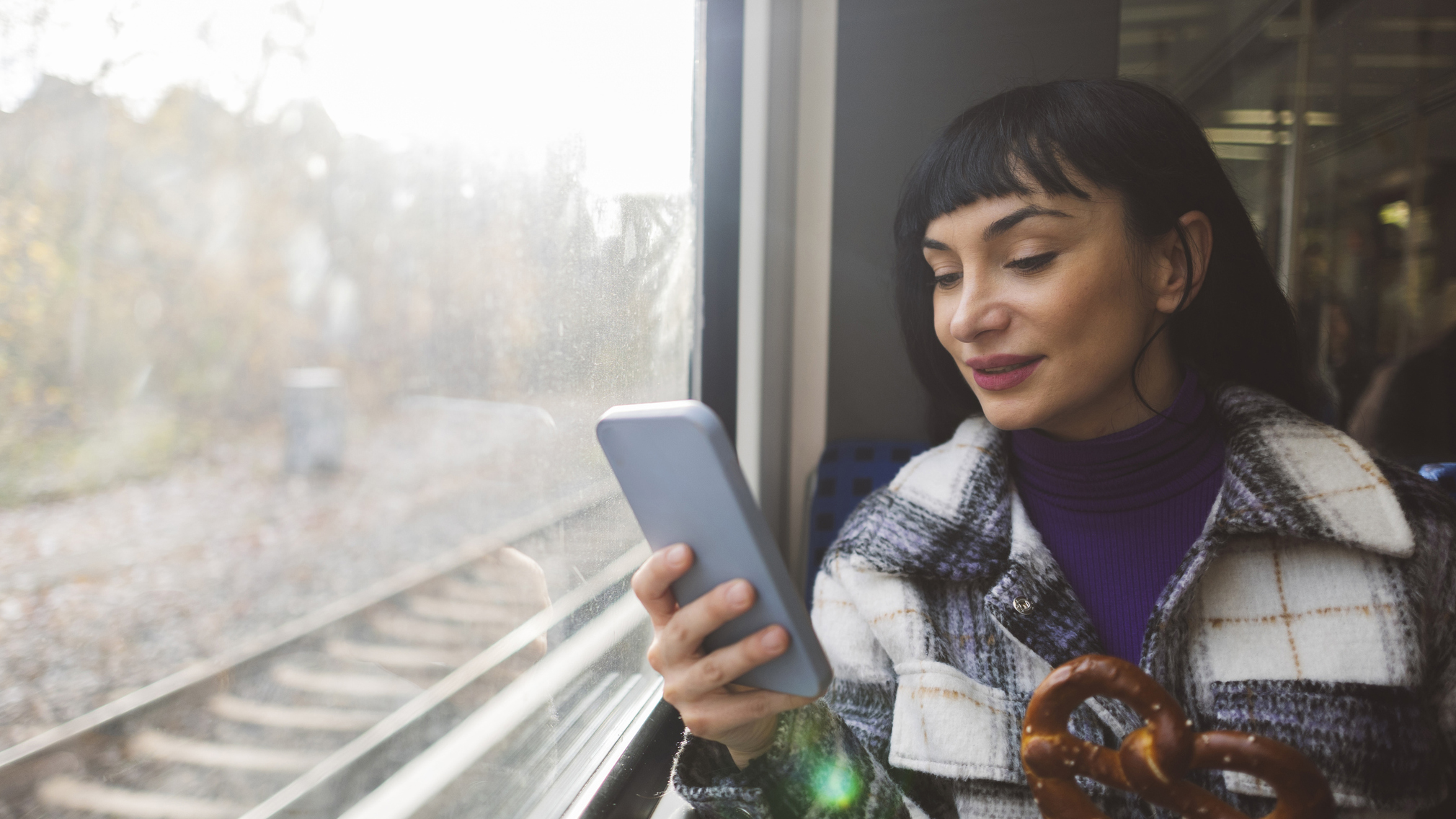 A woman holds a pretzel and uses a smart phone while on the train