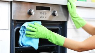 Someone wearing green, rubber gloves cleaning the exterior of an oven