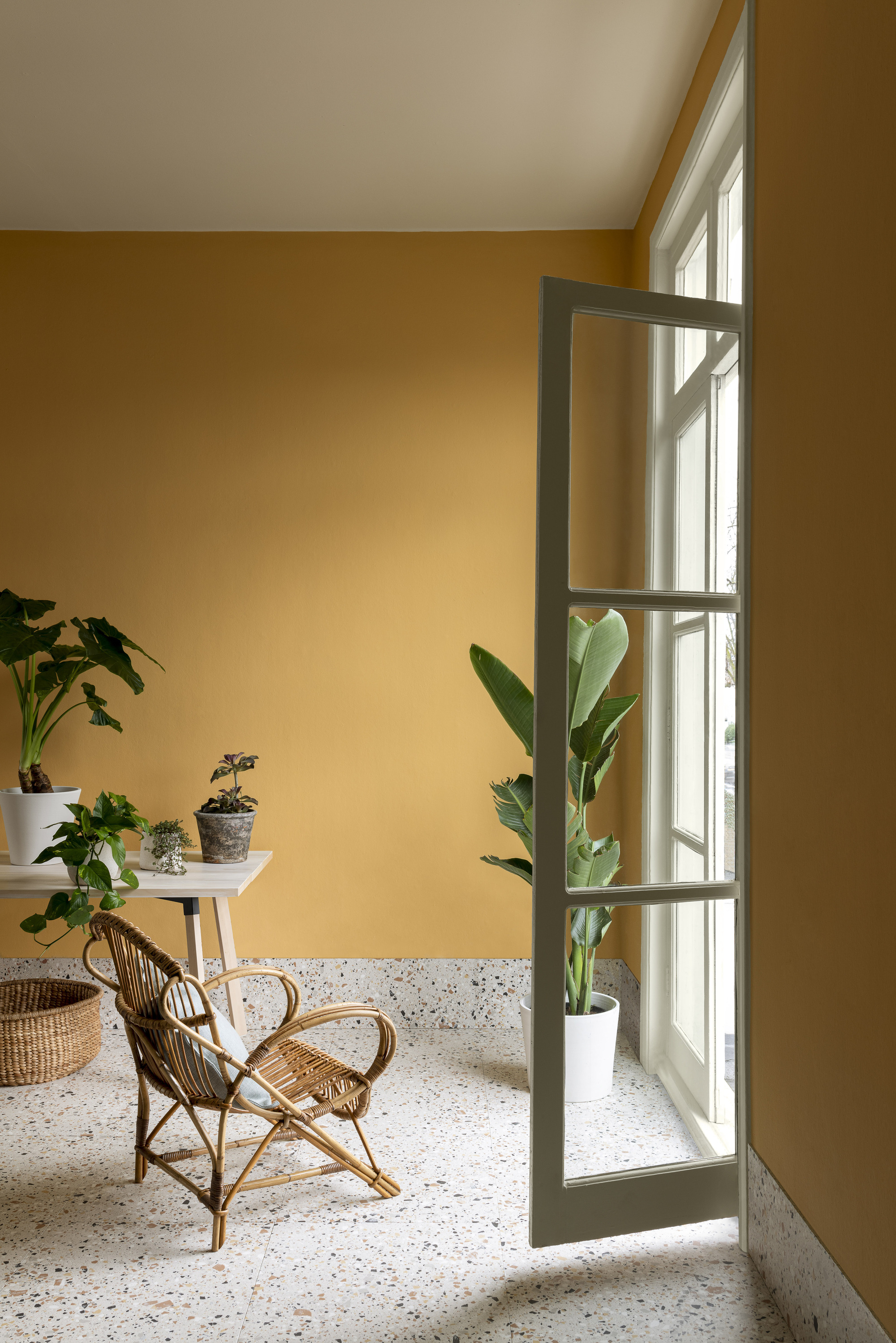 yellow room with terrazzo floor, potted plant, french doors open, and a cane chair with cushion next to a desk covered in pot plants