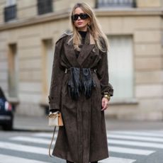  Virginie Conte wears black sunglasses, a dark brown top, a brown suede trench coat, a brown suede skirt, a black leather belt, black leather gloves, gold bracelets, a white leather Kelly Hermes bag, black lace tights with floral details, brown leather Gucci shoes high heels, during a street style fashion photo session, on November 30, 2024 in Paris, France.