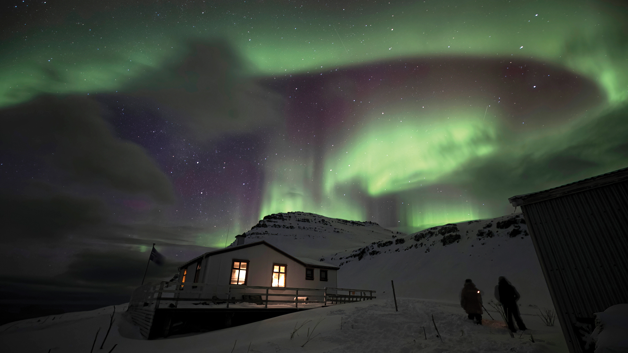 A cabin in snowy terrain under vibrant green and purple northern lights, with two figures walking nearby and stars twinkling above