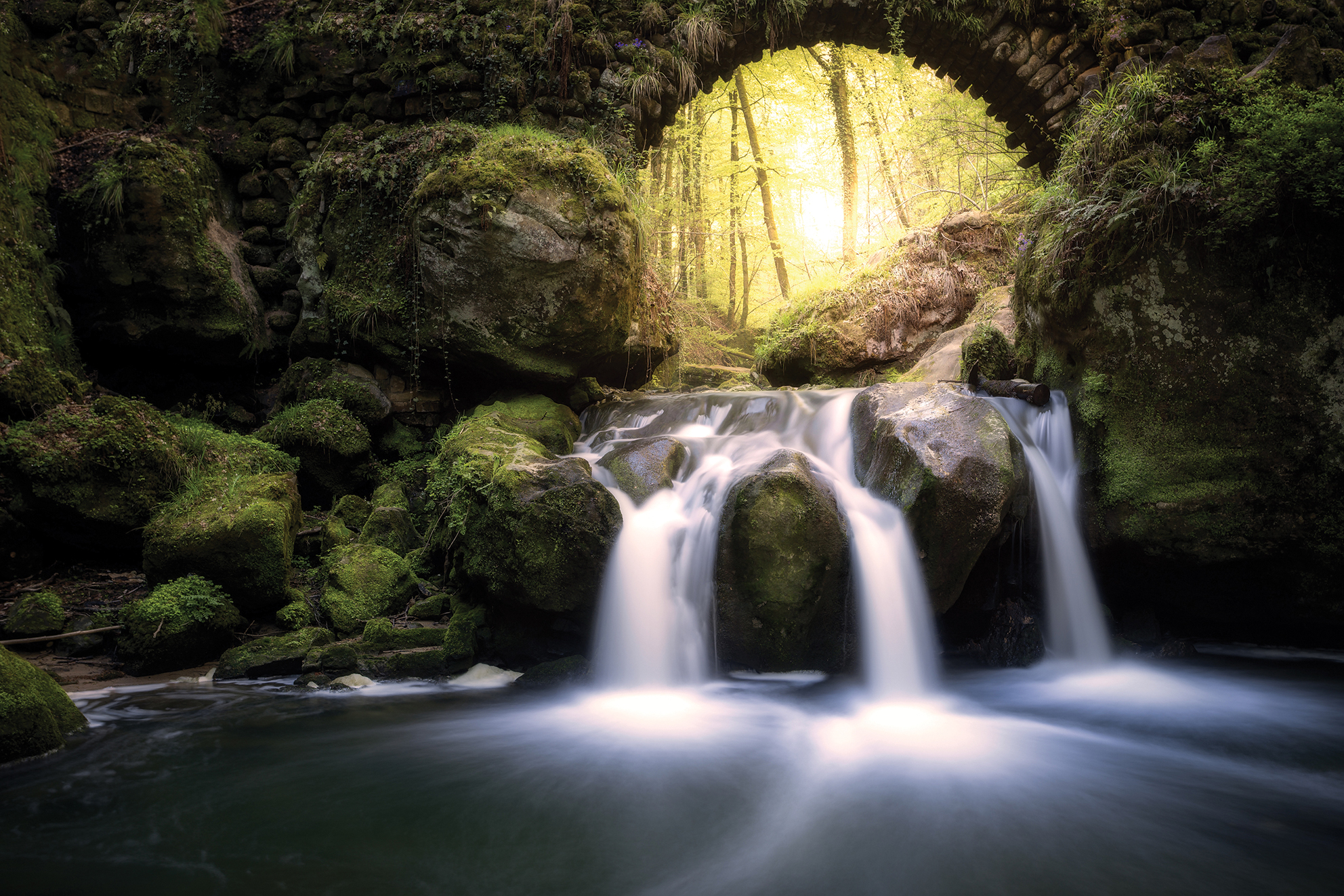 A serene waterfall flows over moss-covered rocks under an ancient stone arch. Sunlight filters through dense forest