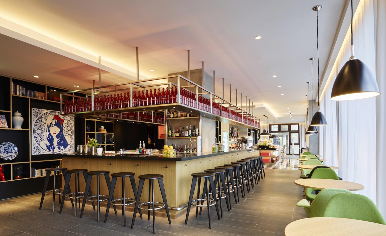 Bar with wooden counter and brass bulkhead frames filled with red bottles, brown stools and round tables to left with lime green chairs
