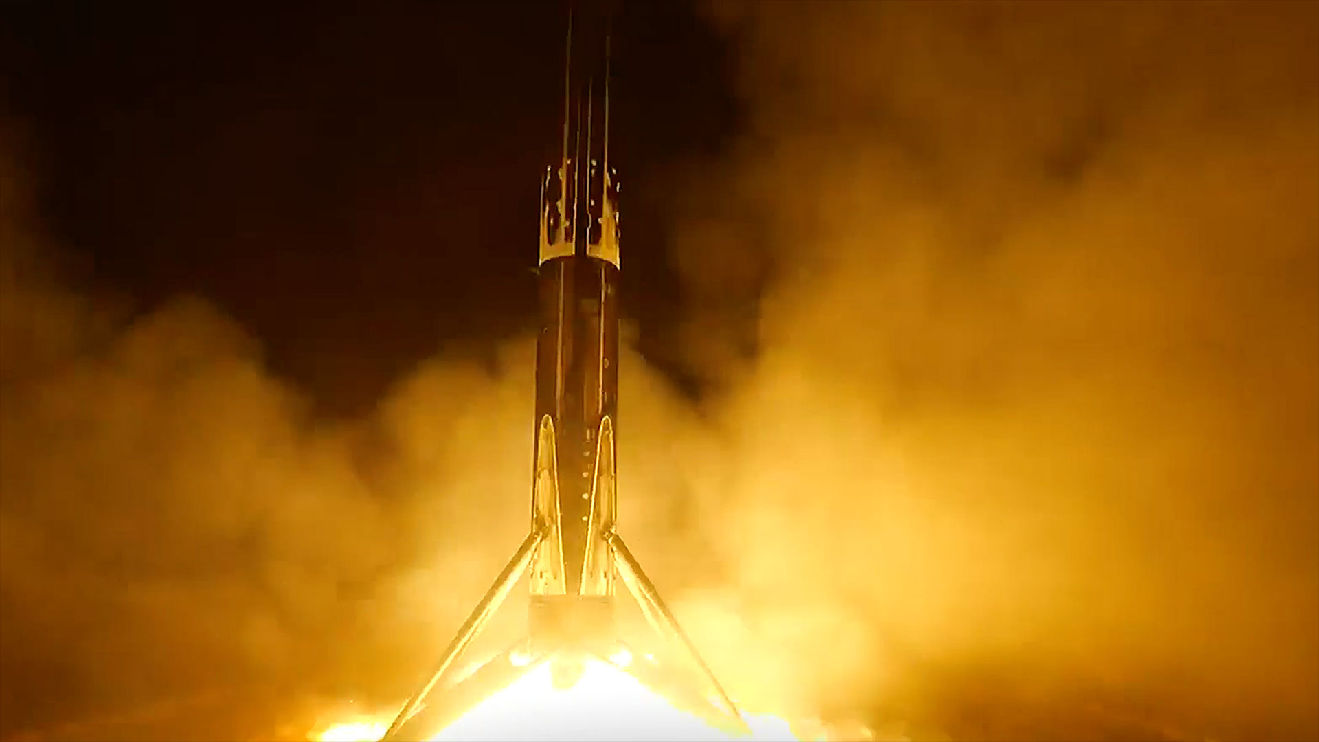 the first stage of a rocket touches down as its engine fires a bright orange flame on a sea-based droneship at night