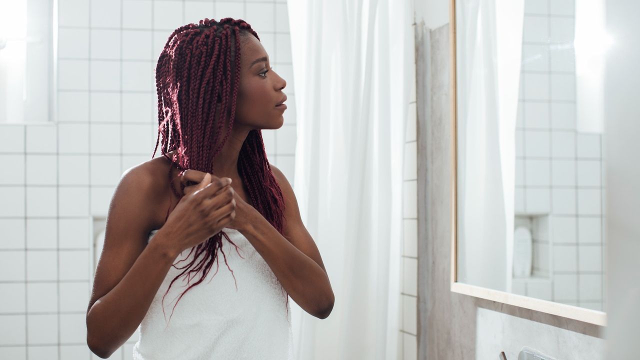 Young woman standing at bathroom and looking at mirror while holding hair.