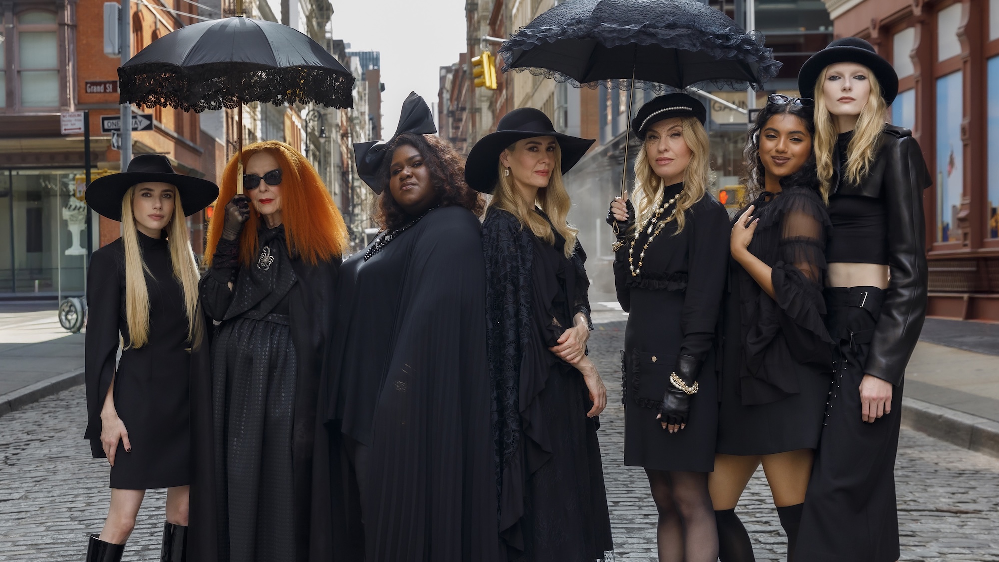 Madison, Myrtle, Queenie, Cordelia, Coco, Ophelia, and Morwenna dressed in black outfits, some with hats, some holding umbrellas, standing in a New Orleans street in American Horror Story 13