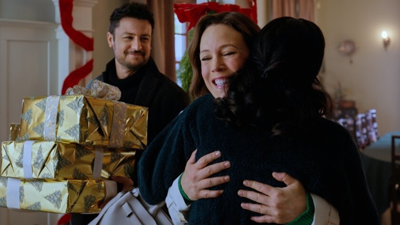 Jake (Tyler Hynes) enters a festive home holding a heap of gold-wrapped presents, looking on while a beaming Ella (Erin Krakow) is reunited with a loved one in Christmas Above the Clouds. 