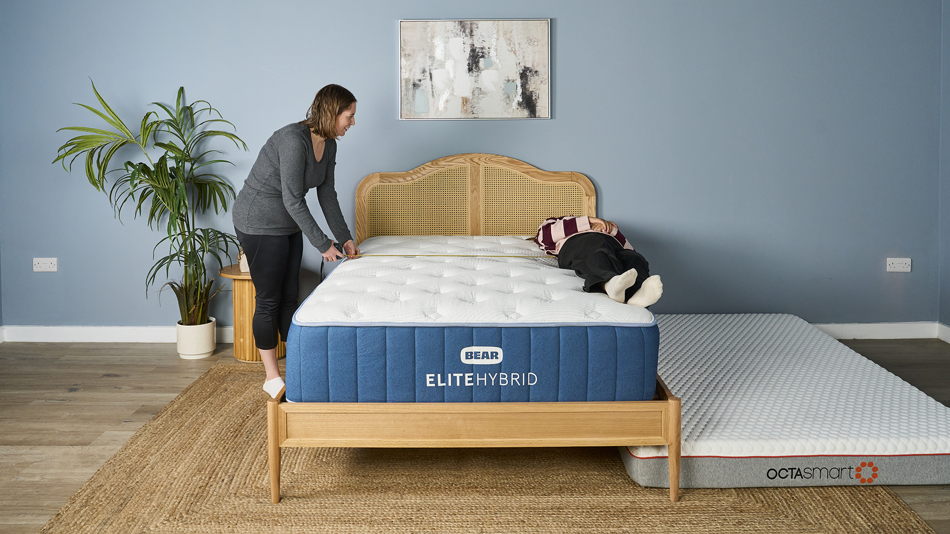A person in a gray top and black pants is pulling up the edge of a mattress protector or sheet on the Bear Elite Hybrid mattress, while a second person is lying near the edge. This shows a test of how well the mattress edge retains its structure and loft when manipulated. The Octasmart mattress lies on the floor next to the bed.