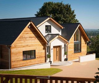 A new build with three gable ends. The central gable, which hosts an integral porch, is clad in white render, with the adjacent gables clad in timber. The house features a driveway and lawned front garden, pictured behind a gate.