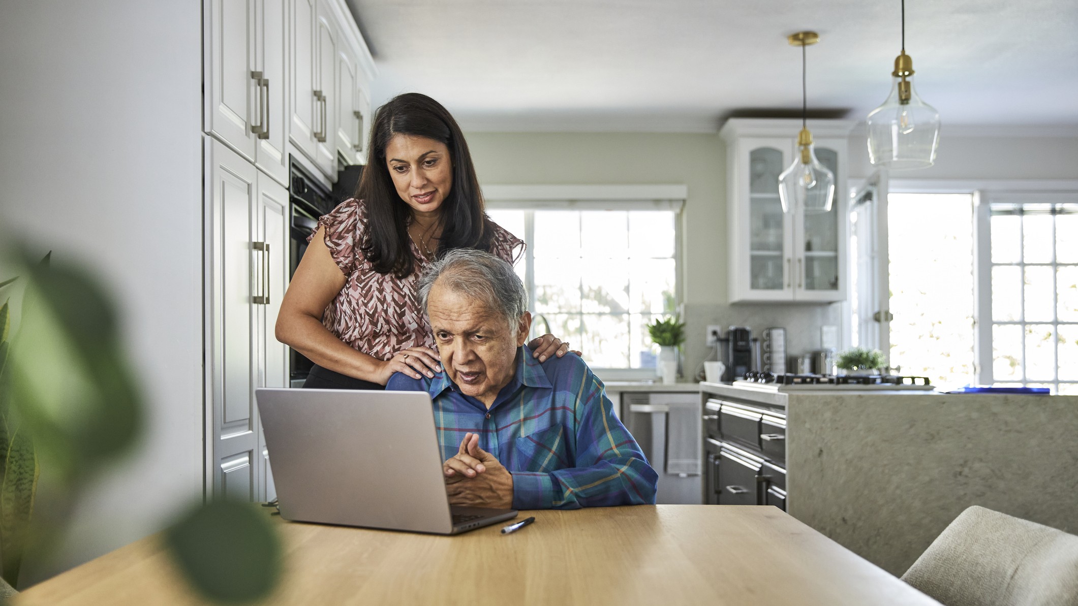 A man sits at a laptop with his adult daughter working with him. 
