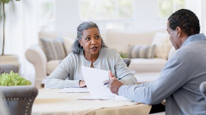 An older woman looks questioning as she talks to a man while sitting together at a table and looking at paperwork.