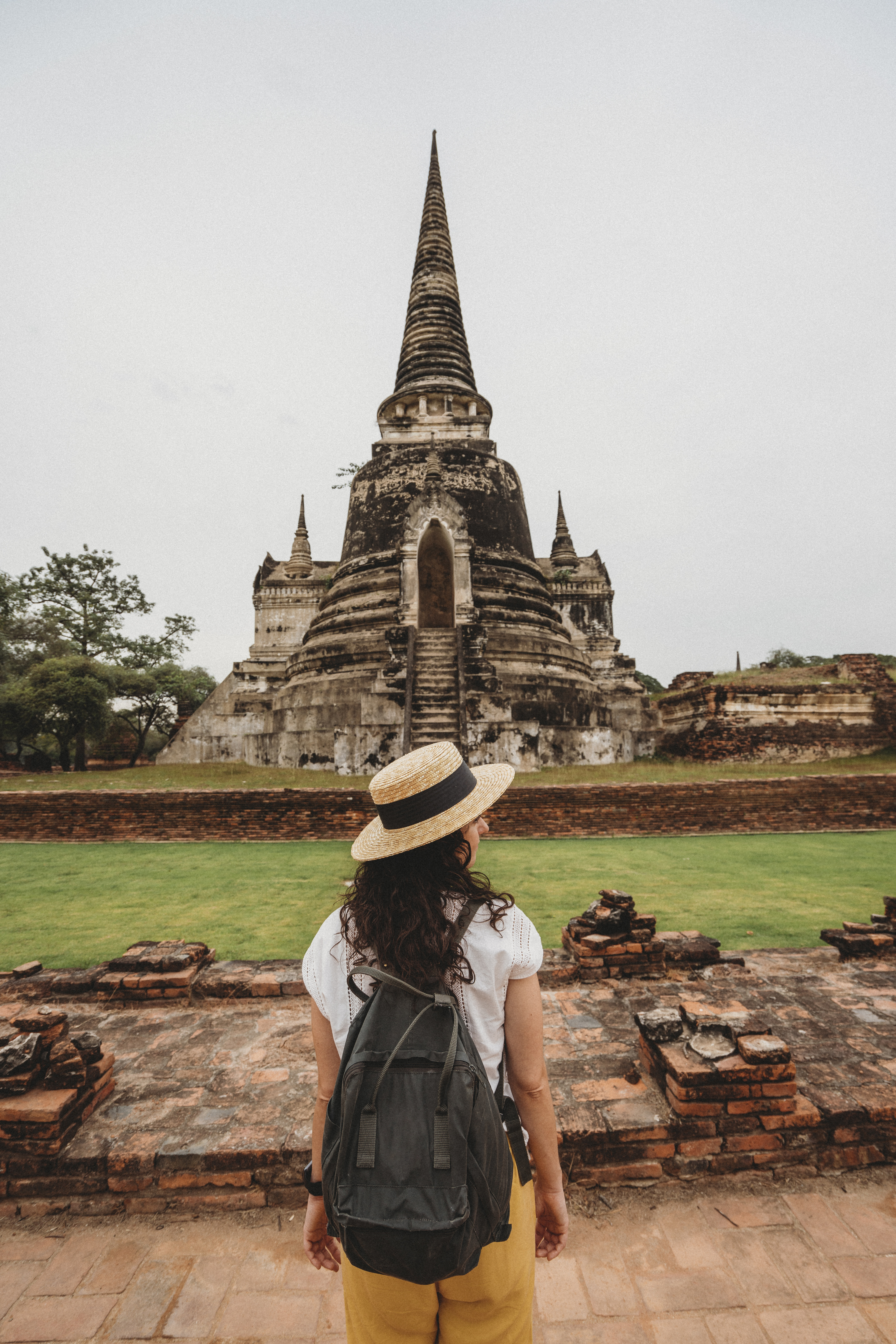 Young woman in a straw hat and wearing a black backpack standing infront of the ancient temples in Ayutthaya, Thailand