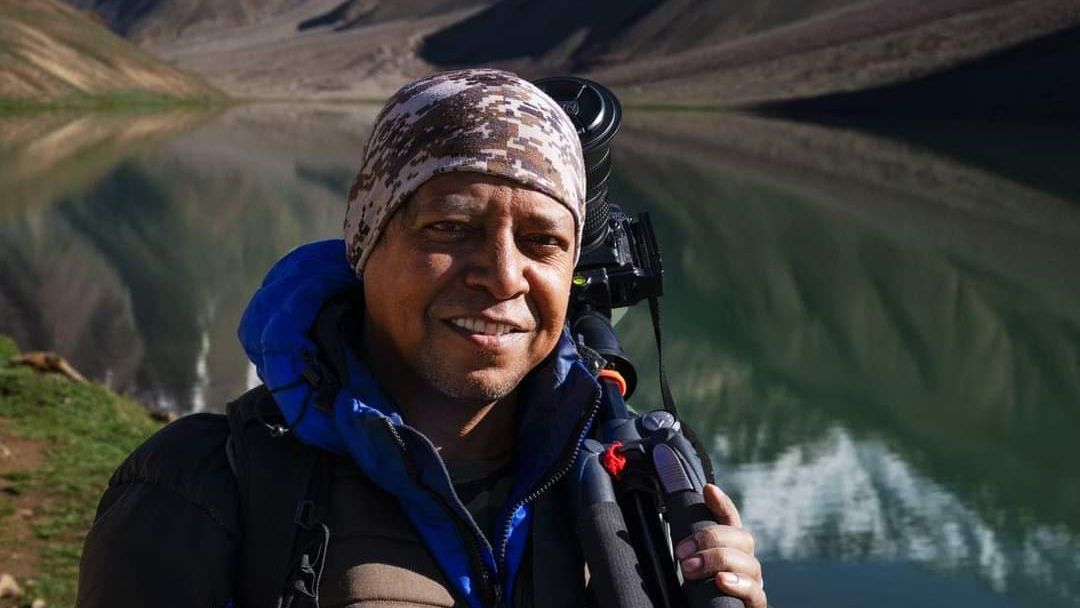A person in outdoor clothing holds a camera, set against a serene mountain lake reflecting the surrounding landscape