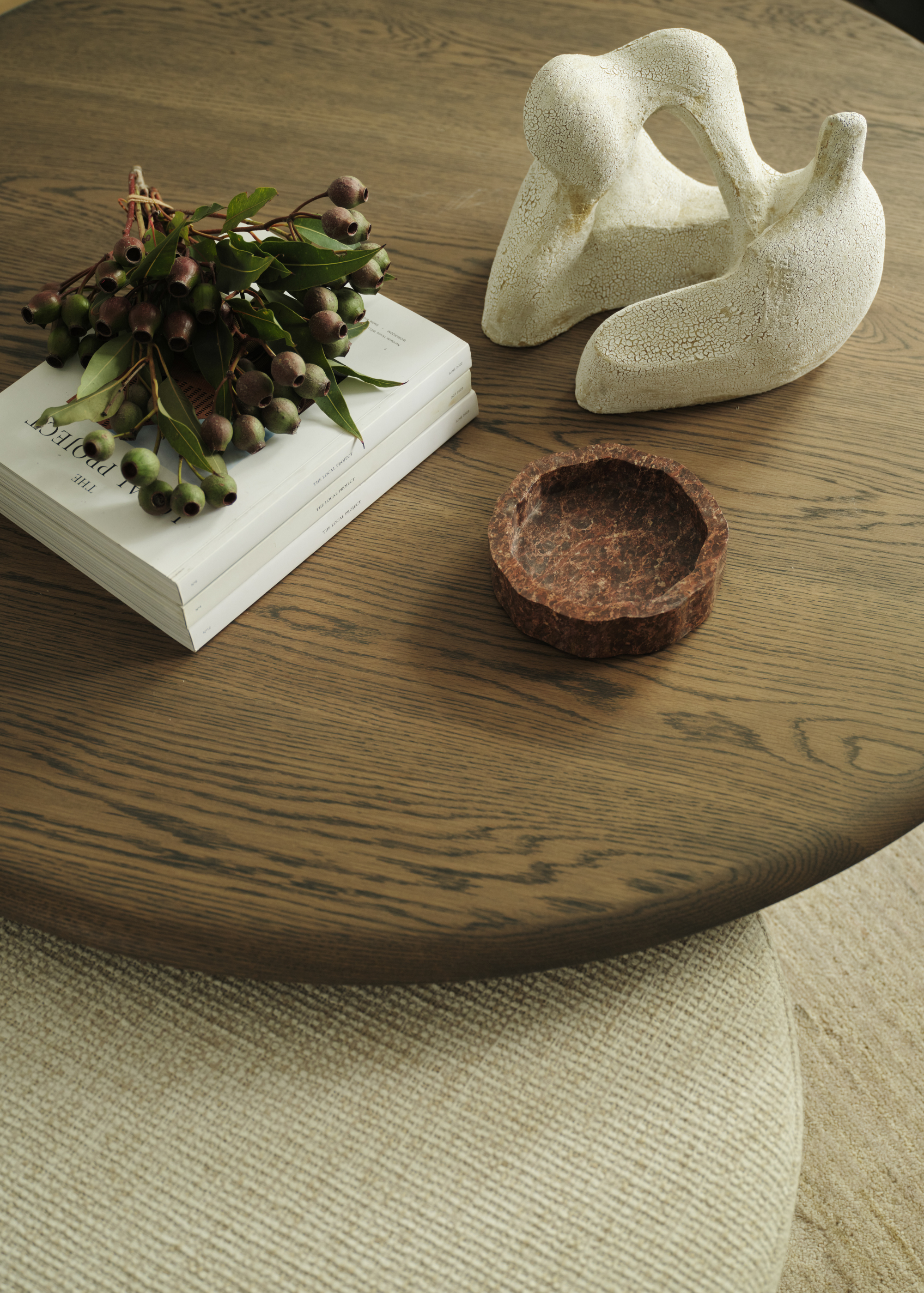 A close-up of a living room coffee table with a branch of olives, a stack of books, a stone catch-all, and a sculpture