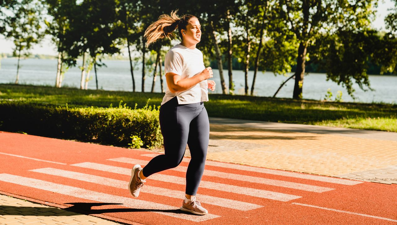 Woman running on running track next to a body of water. She is wearing blue leggings and a white T-shirt and her hair is in a ponytail.