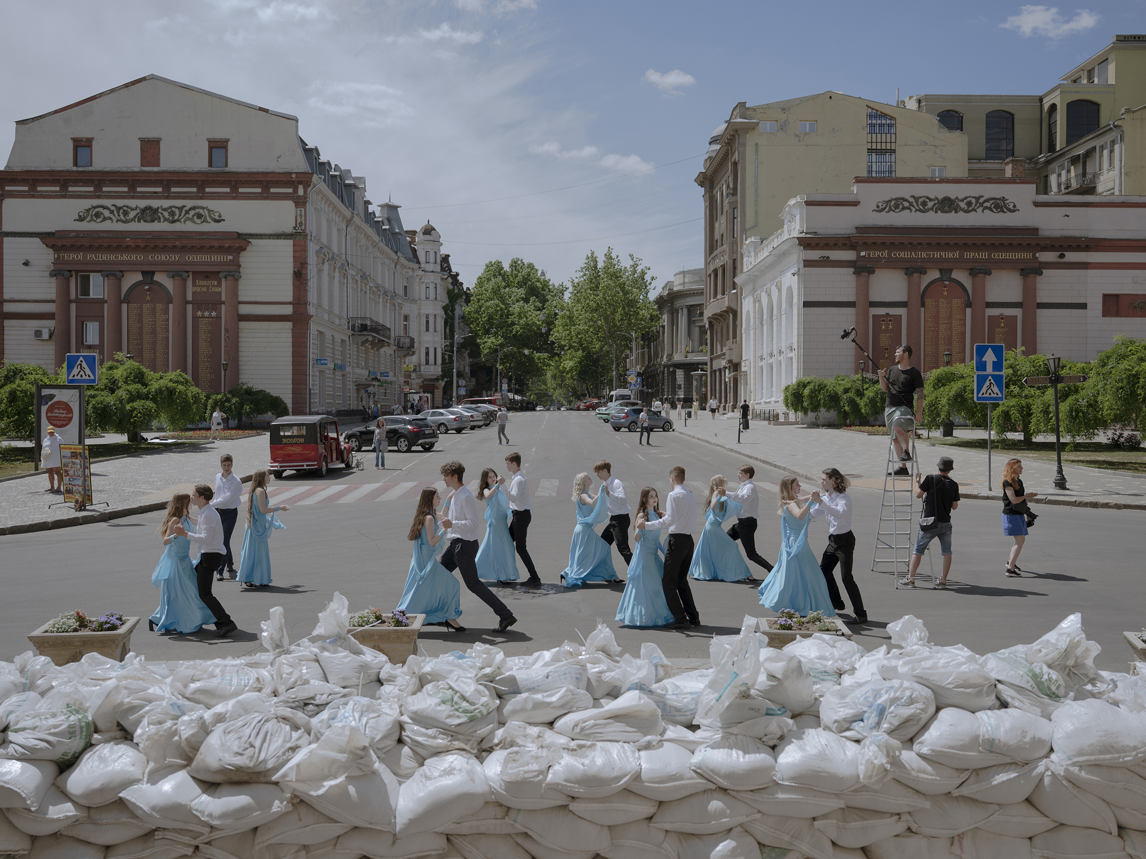 A group of dancers in blue dresses and white shirts perform in a street, bordered by sandbags. Historic buildings set an elegant, contrasting backdrop