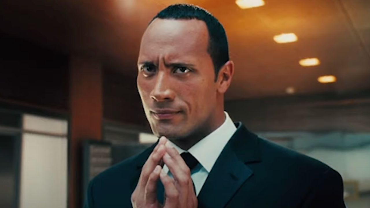 Dwayne Johnson tents his fingers cryptically while smiling in a black suit in Southland Tales.