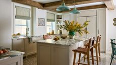 A kitchen with neutral cabinets, white walls, marble counters and a kitchen island with two tan bar stools and wood ceiling beams. 