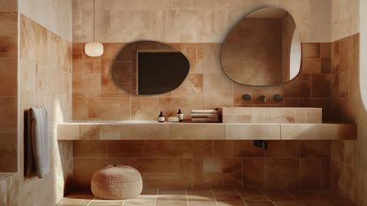 A modern beige bathroom with two irregular shaped mirrors above the sink and joined vanity with a hanging towel and a beige small pouf on the floor with a pendant light hanging above it