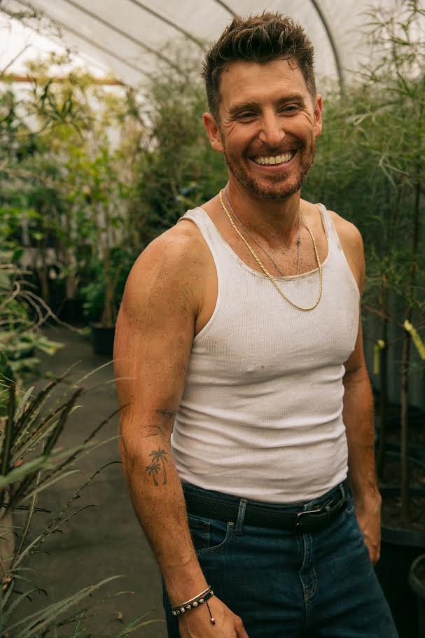 A man with short dark brown hair wearing a white vest and jeans with a black belt and a gold and silver necklace is smiling looking away from the camera standing amongst lots of plants