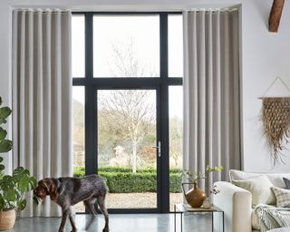 Calm living room with hemp, clay hued floor length drapes framing glass door to rural outdoors.