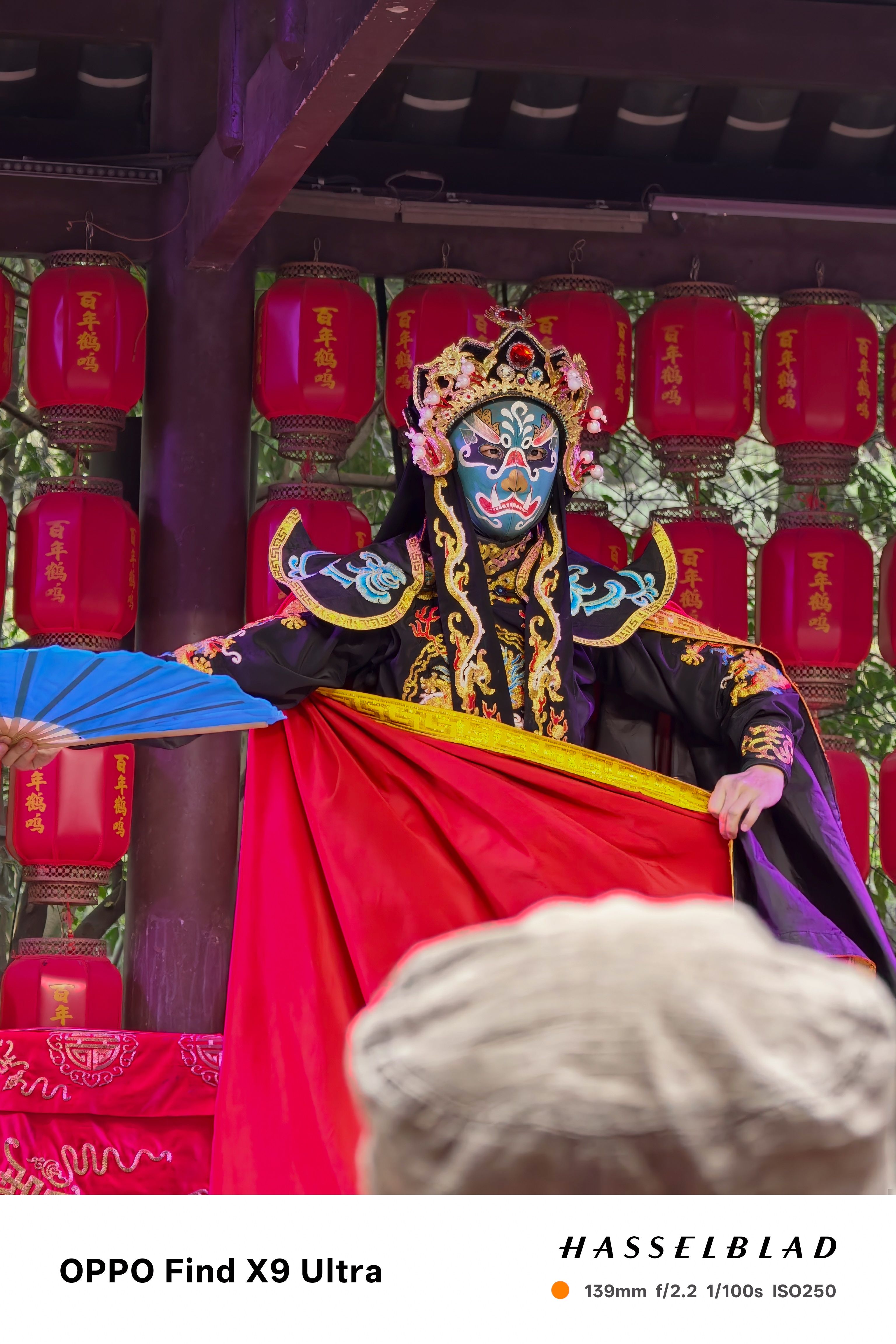 Performer in ornate costume and painted mask, framed by red lanterns on an outdoor stage