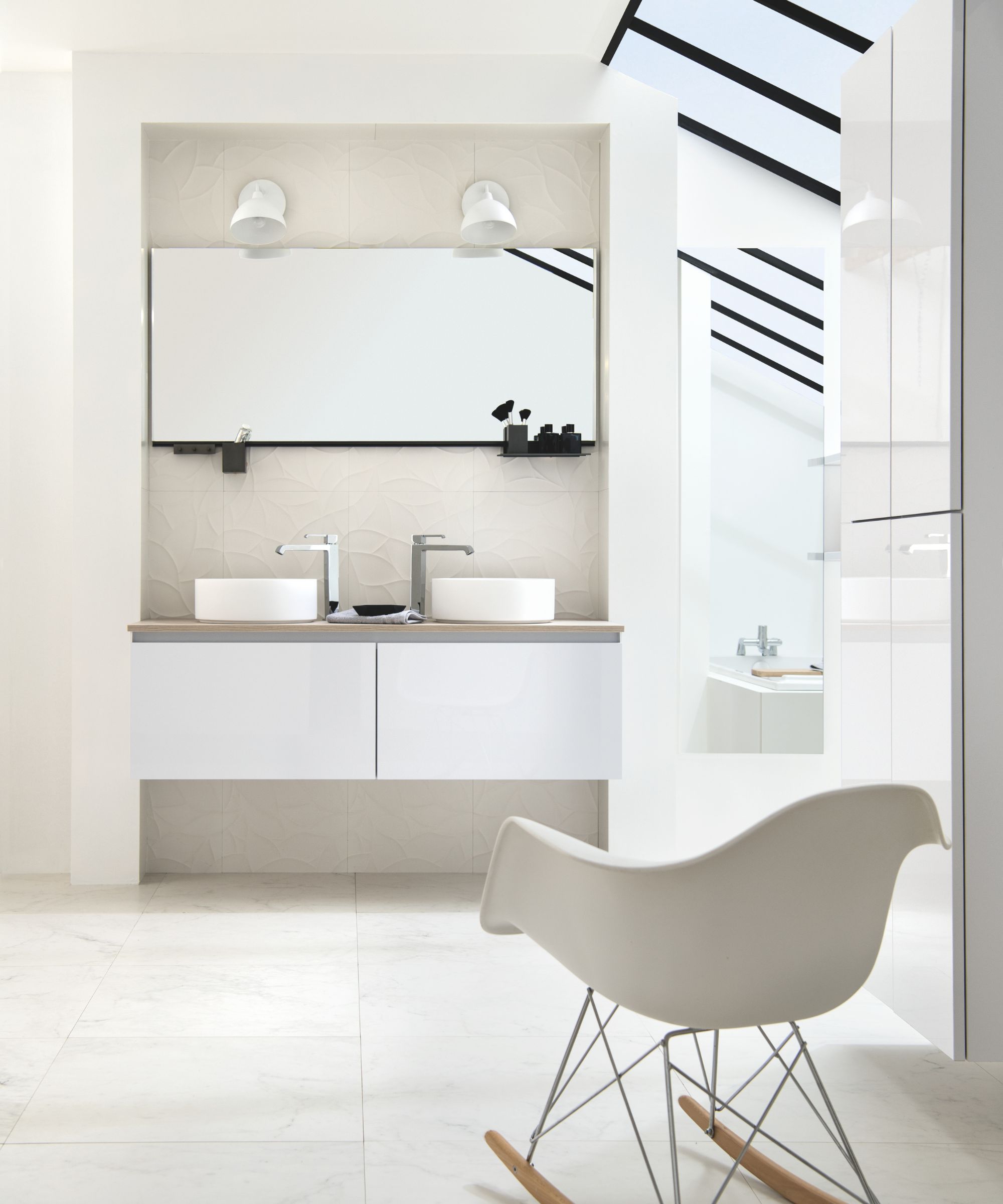A bright, all-white Scandinavian bathroom with large-format stone-effect floor tiles and a subtle textured tile splashback. A white gloss floating vanity unit with an oak worktop holds two white round vessel basins with chrome taps, below a wide frameless mirror with white wall spotlights.