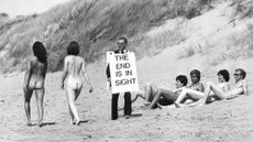 Black and white photo of an evangelist with sandwich board reading THE END IS IN SIGHT walks past naked bathers on a beach in Lancashire in 1979