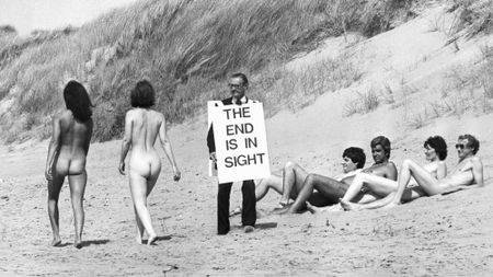 Black and white photo of an evangelist with sandwich board reading THE END IS IN SIGHT walks past naked bathers on a beach in Lancashire in 1979