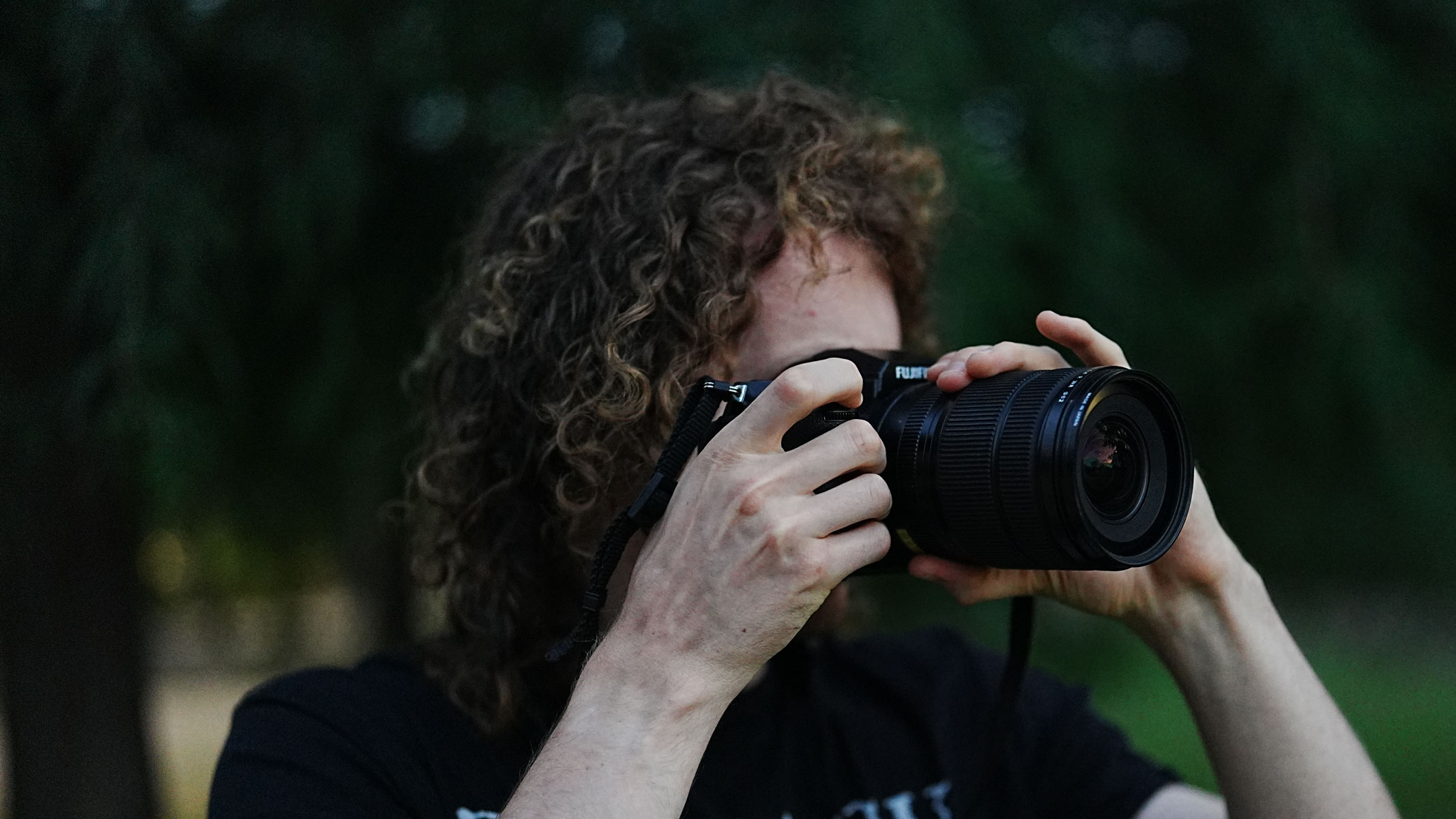 A man with long hair looks through the viewfinder on the Fujifilm GFX100S II, with trees in the background.