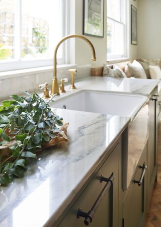 Close up of kitchen sink and veined sideboards with gold taps. There is an out of focus sofa in the background.