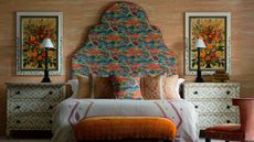 A colorful bedroom with a large bed with oversized patterned headboard, white bedding, assorted colorful cushions, and an orange ottoman at the end of the bed. To either side are white and chest of drawers covered in green geometric patterns, with brown lamps with small white lampshades on top. Behind either lamp are large floral artworks in white frames, and the wall is wallpapered a pale burnt orange with white detailing.