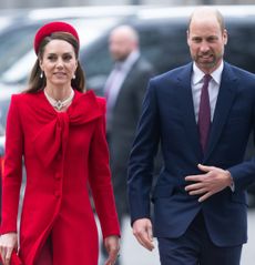 Kate Middleton wearing a red coat and hat walking next to Prince WIlliam wearing a blue suit