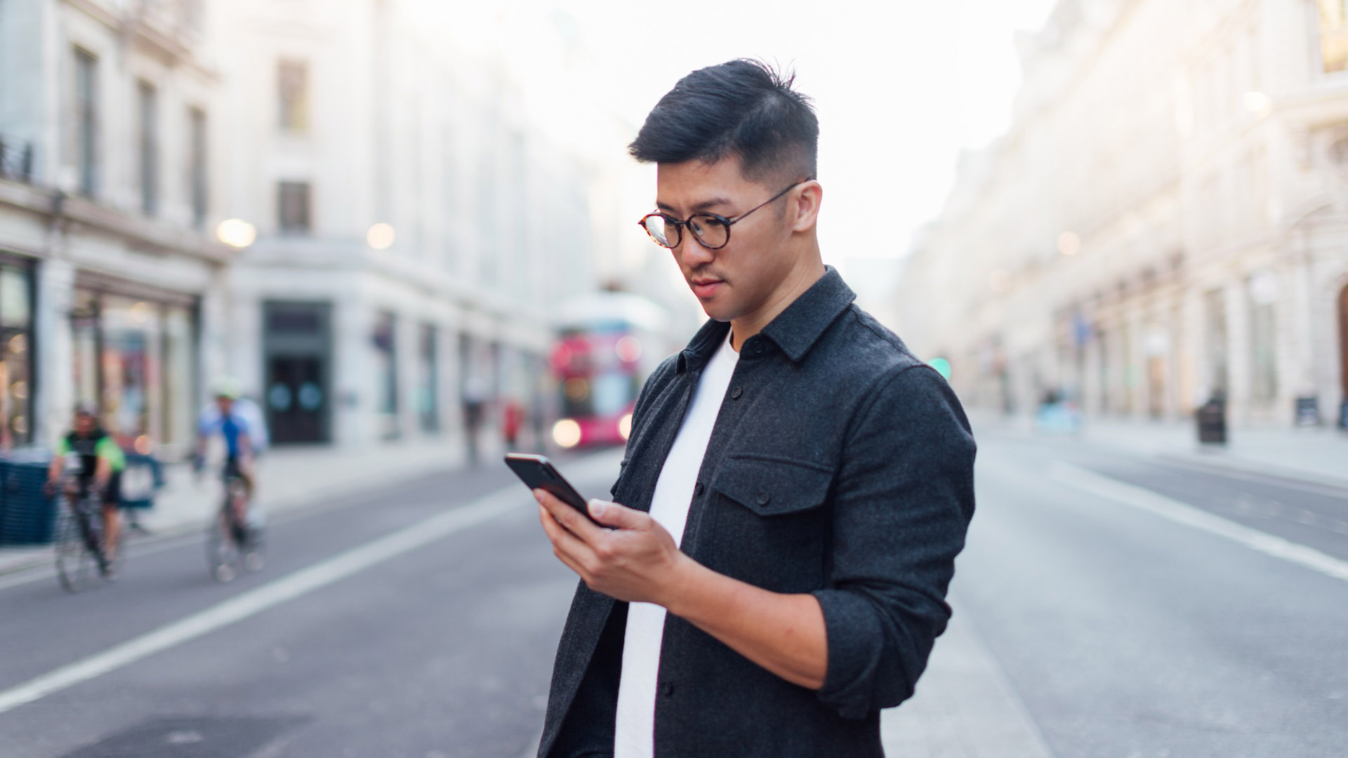 Medium shot of young Asian man checking text messages on smartphone while waiting for taxi, standing on the street in a big city.