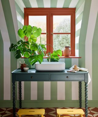 green and white striped wallpaper on wall with blue desk in front of window with red window frame, houseplant on desk