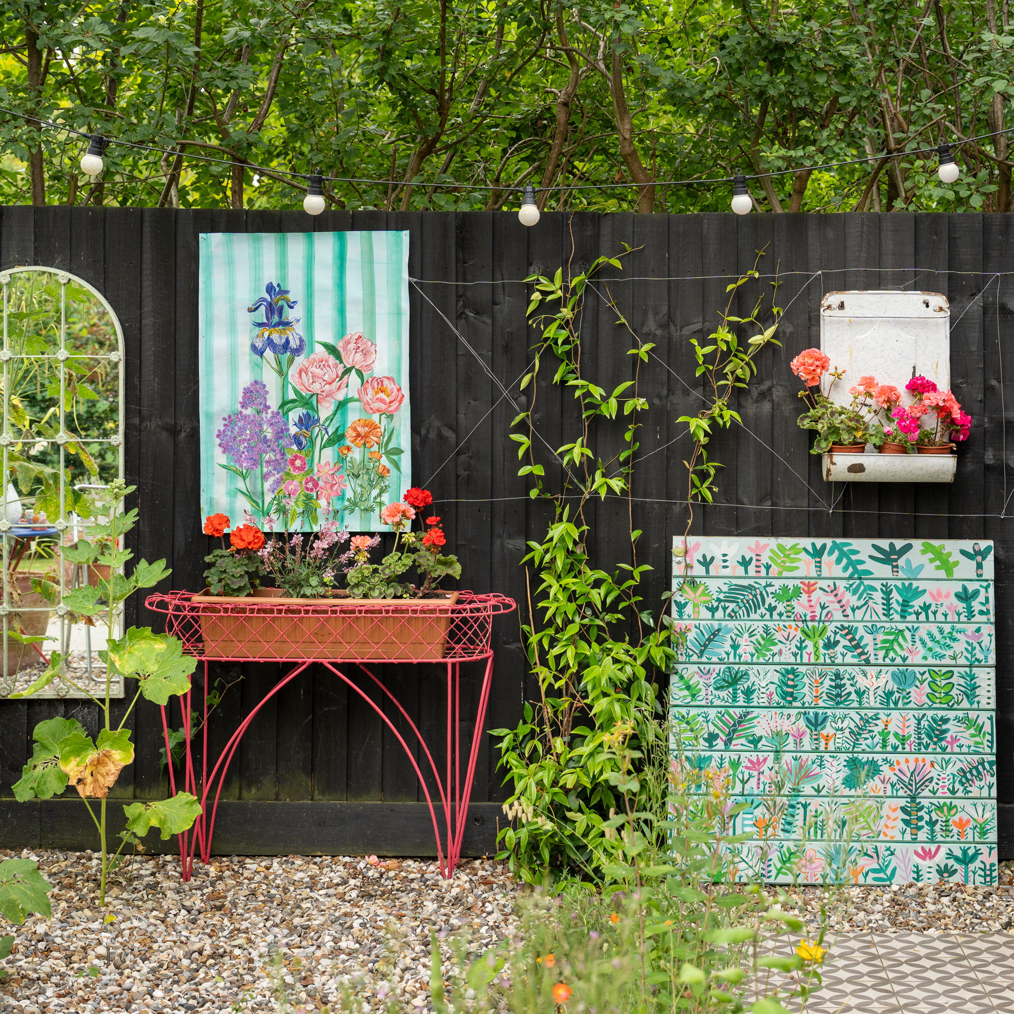 a black painted fence with outdoor artwork a bright red table, arched mirror and bright red geraniums